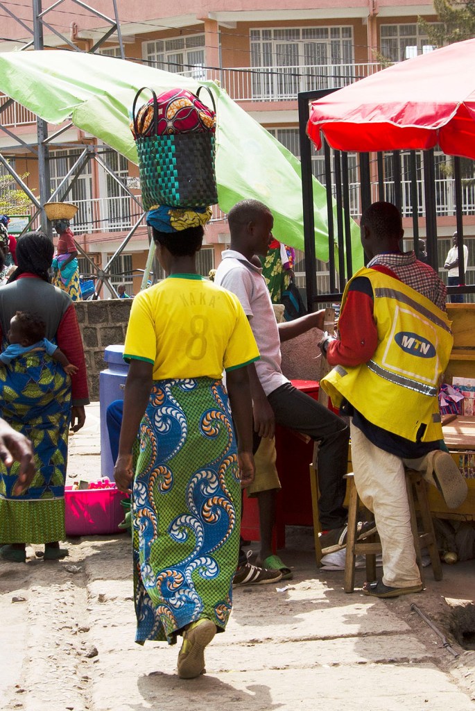 Woman in blue and green printed traditional wraparound attire with a plastic woven bag balanced on her head.